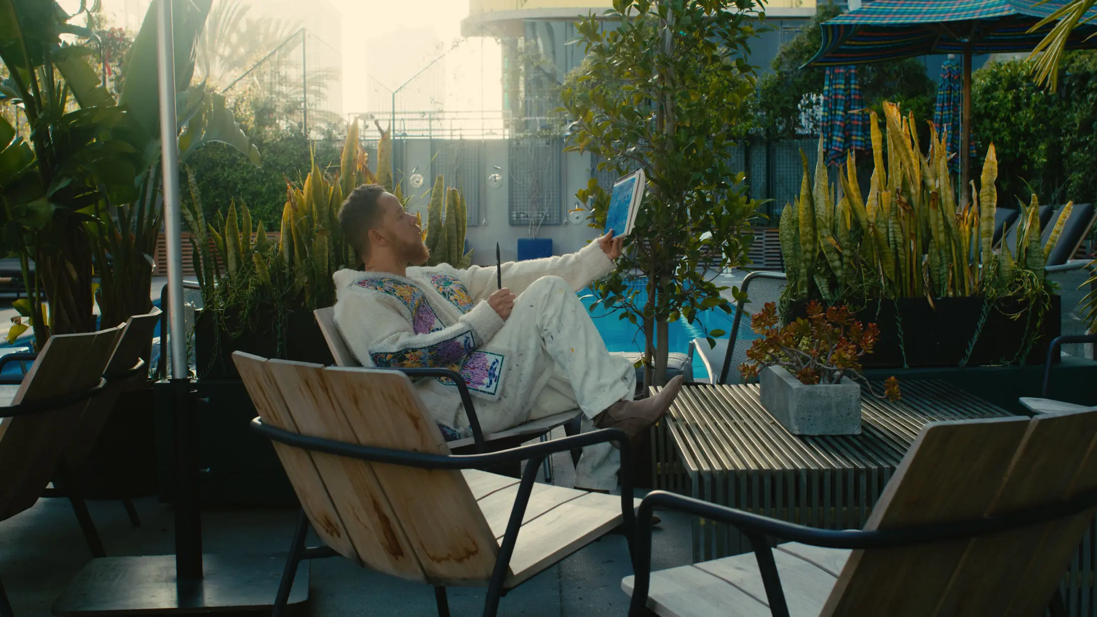 A man in white attire relaxes on a chair while sketching diagrams in a notebook.