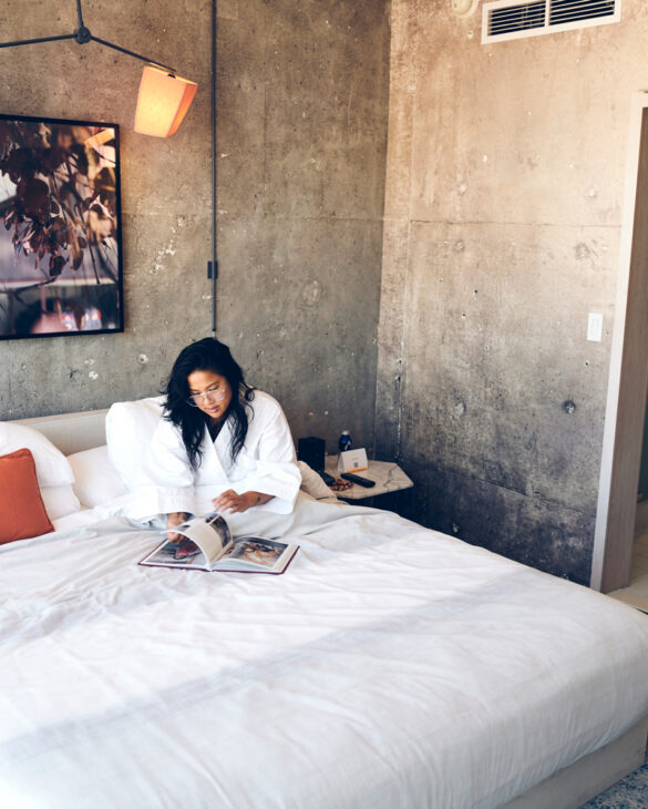 A woman in a bathrobe sits on her bed while reading a magazine.