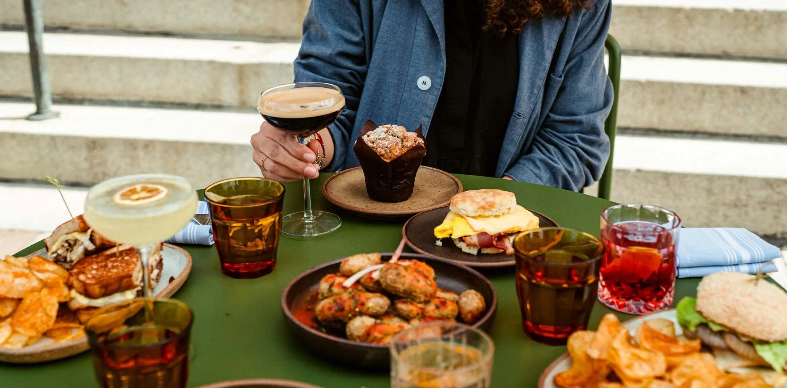 A person sits a patio table covered with dishes of food and drinks