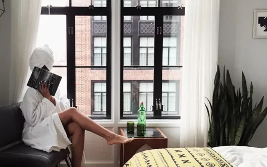 Picture of a women in bath robe sitting on the chair by hiding her face from a book and along with plant pot and drink kept on table