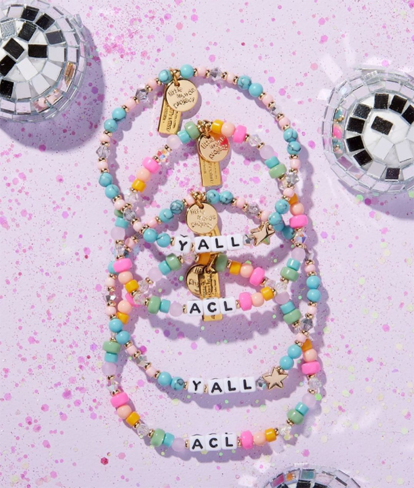 Four colorful beaded bracelets with "YALL" and "ACL" letters loosely stacked on a glittery purple background next to two disco balls