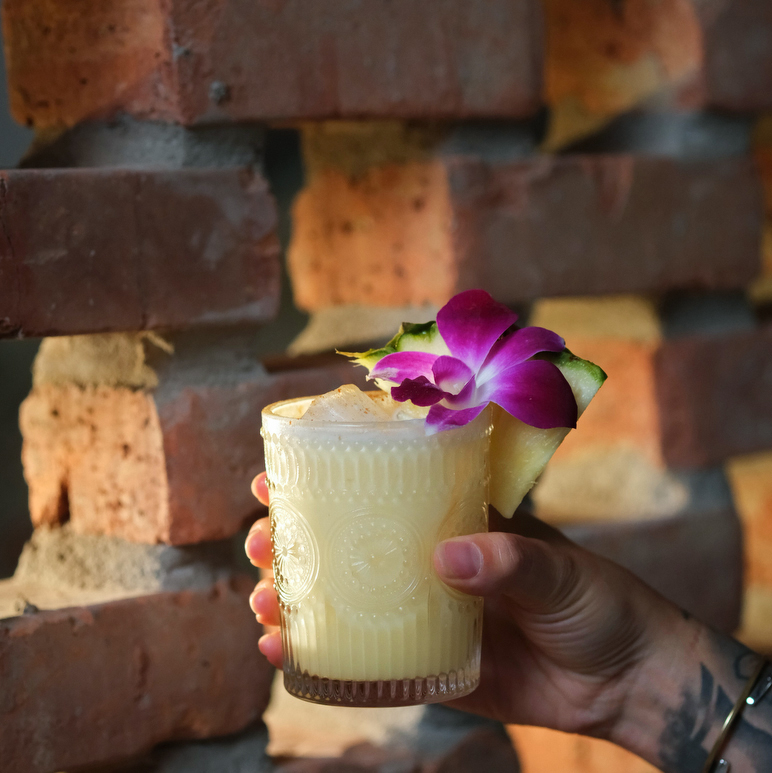 Cantero - A hand holding a glass of cocktail in front of a brick wall