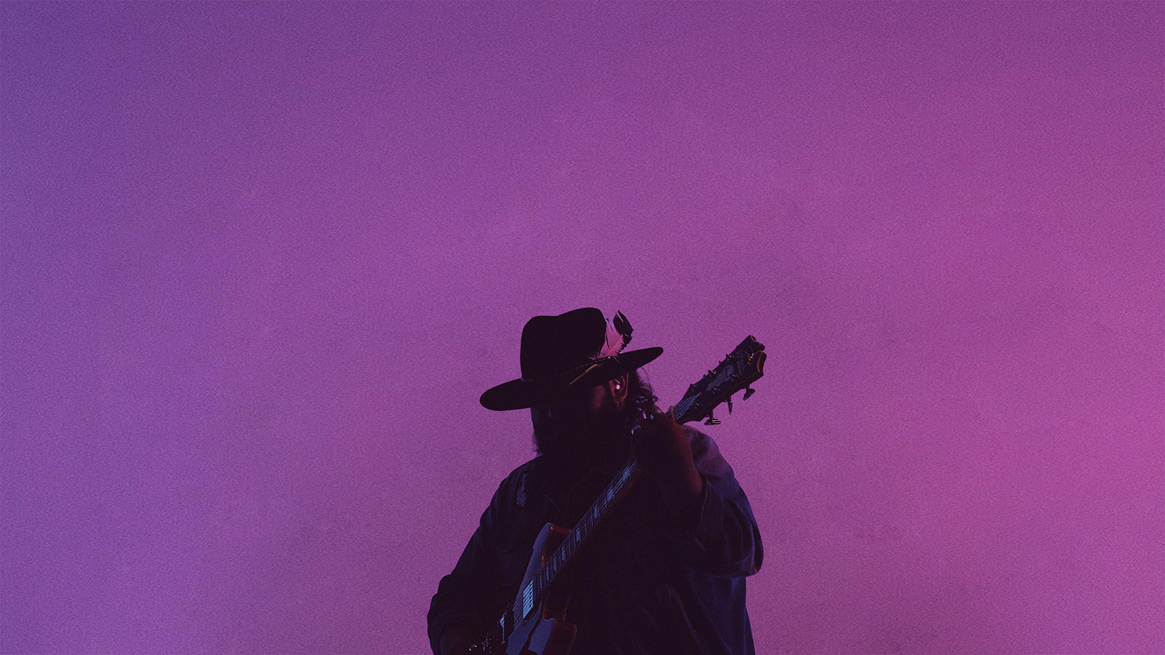 A person wearing cowboy hat holding a guitar in front of a purple background